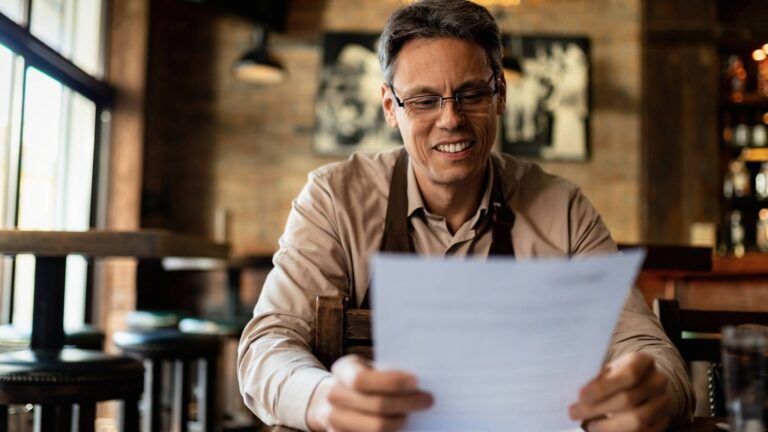 Un hombre con gafas lee un periódico en un restaurante, mientras disfruta de su comida y ambiente acogedor.
