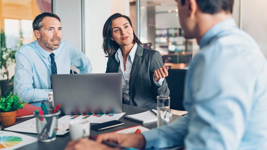 Tres personas de negocios sentadas en una mesa, trabajando juntas en un laptop.