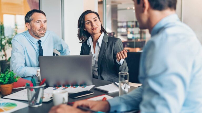 Tres personas de negocios sentadas en una mesa, trabajando juntas en un laptop.