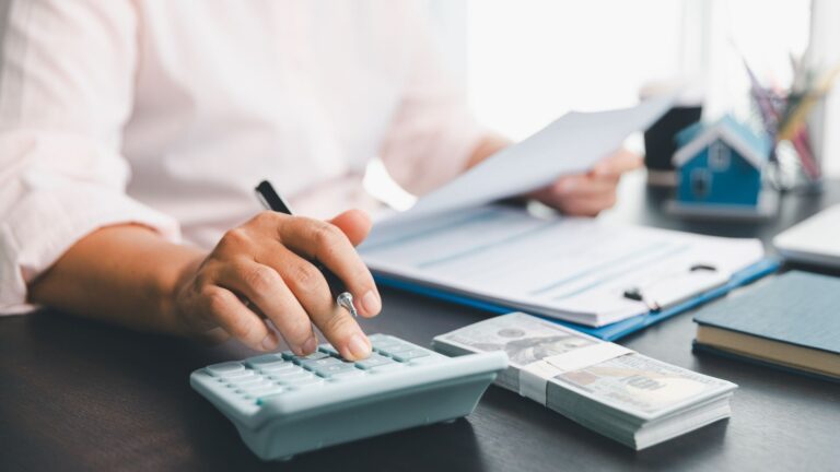 Mano de mujer escribiendo en una calculadora mientras sostiene dinero, evaluando gastos o ingresos.