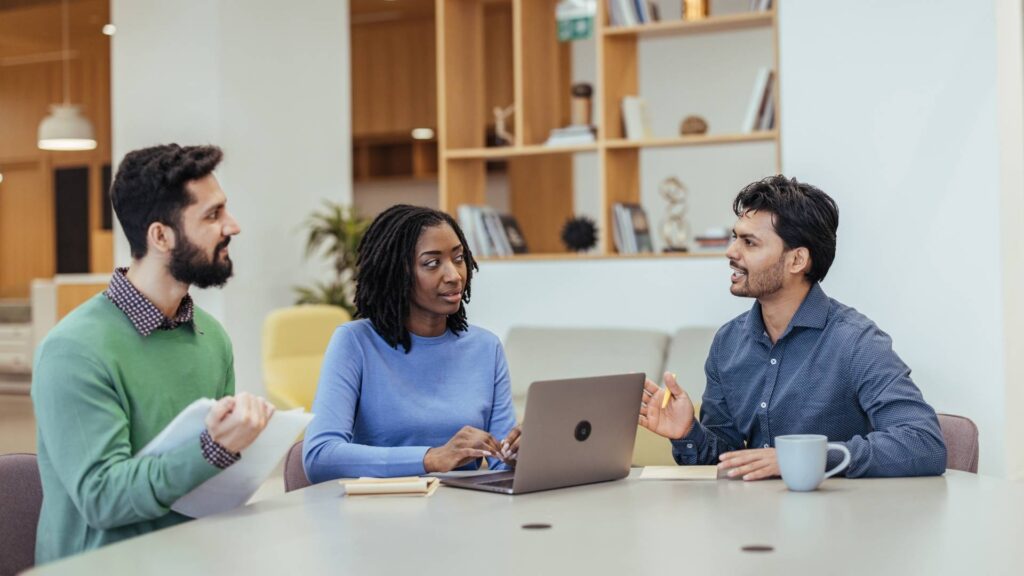 Tres personas sentadas alrededor de una mesa, cada una con un portátil, colaborando en una evaluación.