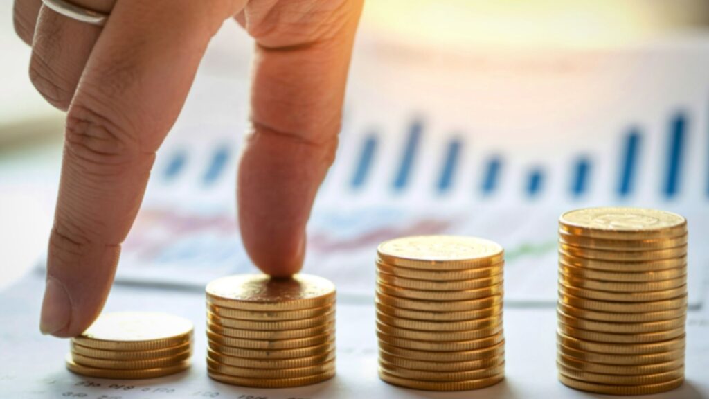 Mano simulando subir escalones sobre pilas de monedas de oro frente a un gráfico financiero