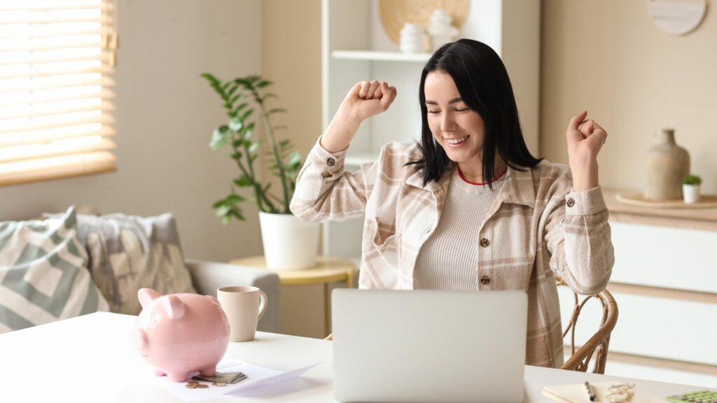 Mujer emprendedora celebrando el éxito financiero frente a su laptop con una alcancía de cerdo.