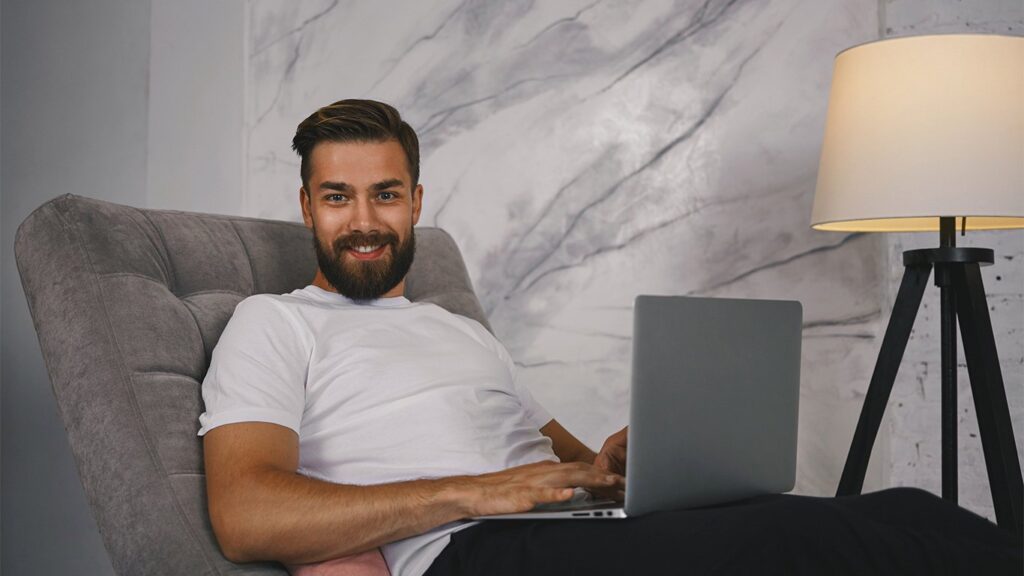 Un hombre sonriente con barba está sentado en una silla cómoda usando una computadora portátil