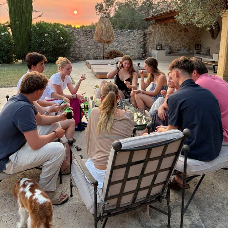 Un grupo de amigos se sienta alrededor de una mesa en un patio al atardecer, disfrutando de bebidas y charlando.