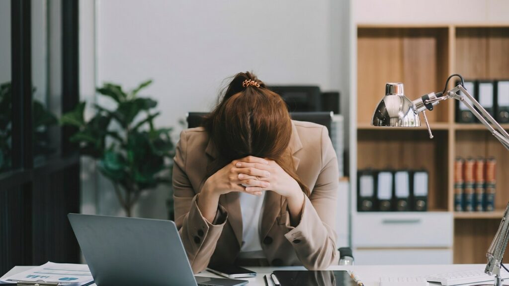 Mujer en oficina, con chaqueta beige, agachada sobre el escritorio con la cabeza entre las manos, mostrando agotamiento extremo.