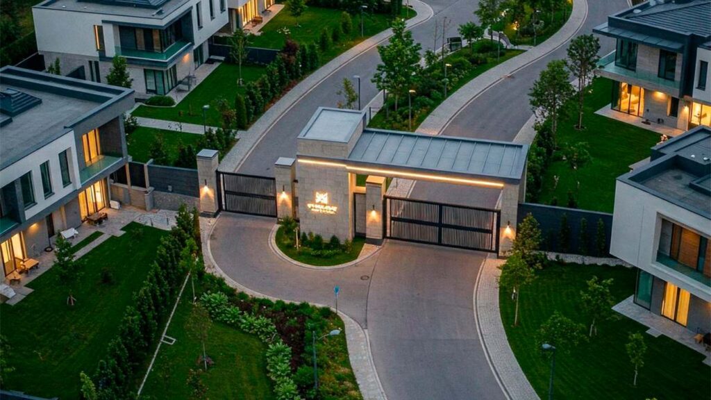 Vista aérea de una lujosa urbanización privada al atardecer, con modernas casas de dos plantas, césped impecablemente cuidado y una entrada tenuemente iluminada con una puerta de seguridad.