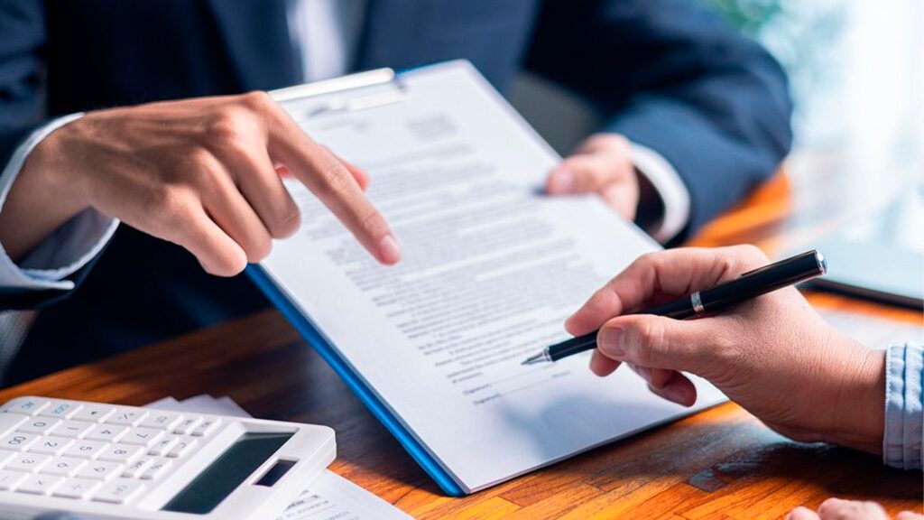 Persona en traje señalando un lugar en un contrato para que otra persona lo firme sobre un escritorio con calculadora.