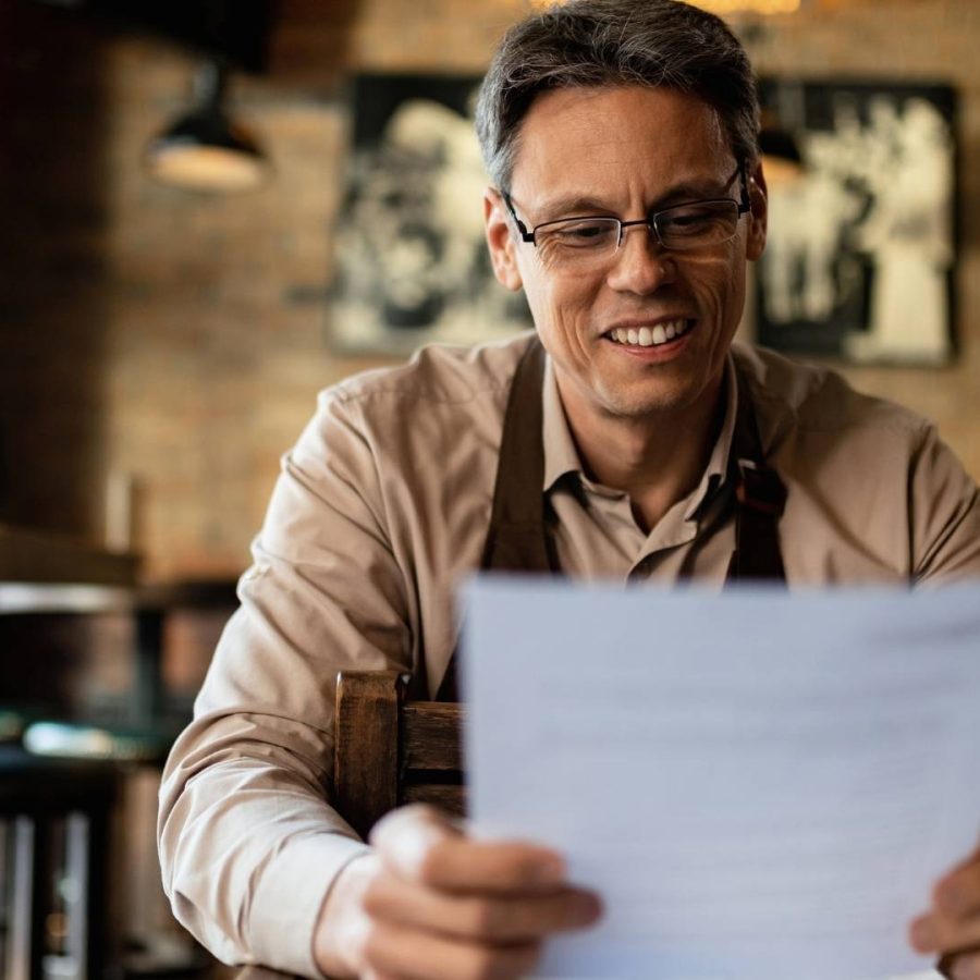 Un hombre con gafas lee un periódico en un restaurante, mientras disfruta de su comida y ambiente acogedor.