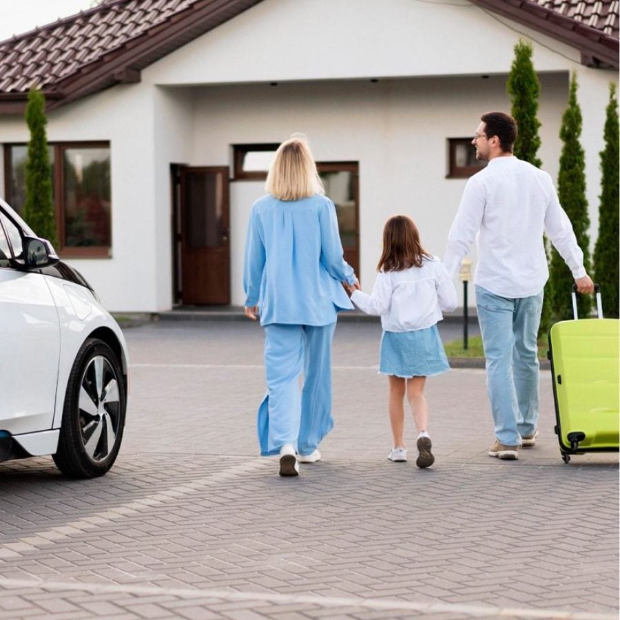 Una familia de tres personas caminando de espaldas hacia una casa blanca moderna, el padre lleva una maleta verde brillante y hay un coche eléctrico estacionado al lado.